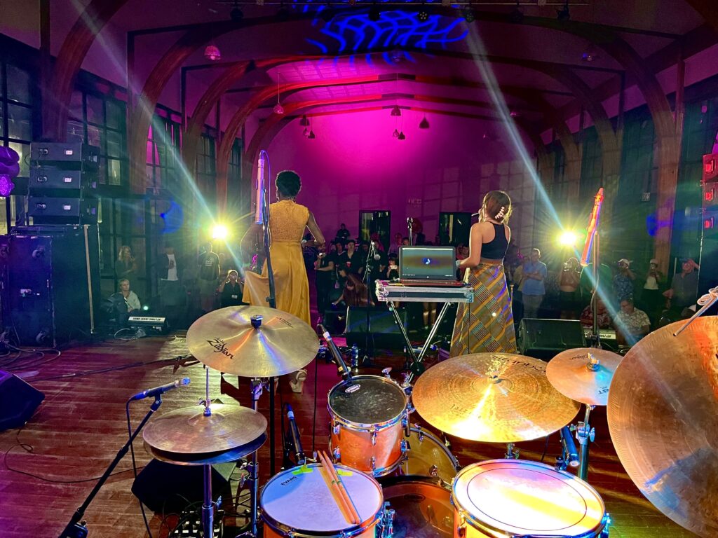 Color photo shot from behind two performers on stage with mics singing to an audience in a venue. In the foreground, a full drum kit can be seen.