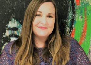 Color photo of board member Jessica Ball. Jessica has long straight brown hair and wears a purple blouse. Jessica is shot from the chest up against a vibrant background.