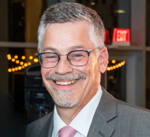 Color headshot of board member John MbW=Ewen. John wears a charcoal grey suit, white shirt, and pink tie. He has greying hair, a grey beard, and wears dark rimmed glasses.