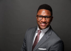 Color photo of board member Nicholas Hawkins. Nick has short dark hair and dark rimmed glasses. Nick is shot from the chest up and wears a charcoal grey suit, white shirt and pocket square, and tie.