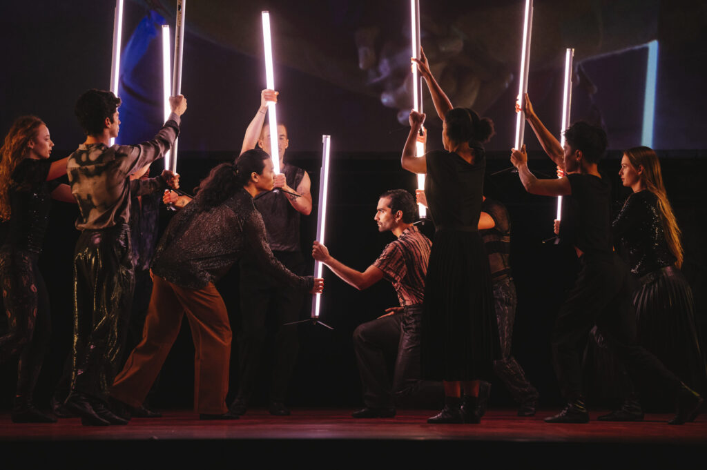 Color image of a large group on stage all holding lit white bars vertically, in the center, two people appear to fight over a single bar.