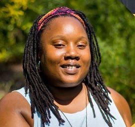 Bekezela Mguni is a Black person with long black braids held off of their face with a bright headband. They wear a light blue tank top and are photographed outside in front of green leaves.