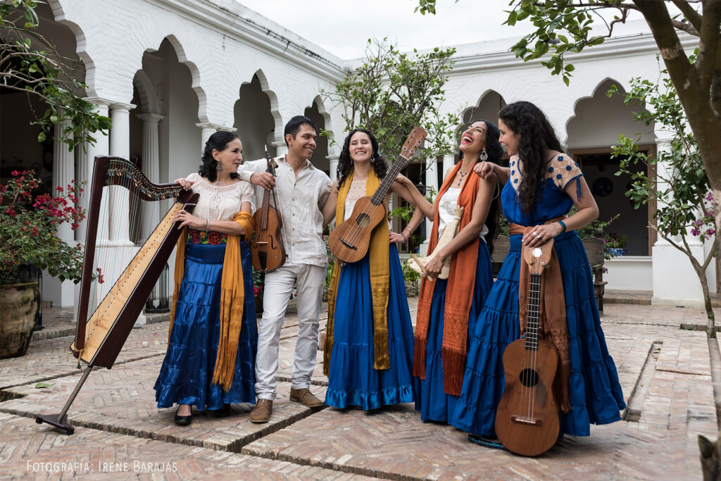 Five musicians in bright blue, yellow, orange, and white traditional dress stand in an ornate courtyard holding traditional instruments.