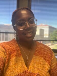 Amaris Mitchell is a Black woman who is wearing gold rimmed glasses and a gold necklace and earrings. Her air has been pulled back and she wears an orange v-necked knit top. She is shot from the chest up in front of a window which overlooks a street and buildings.