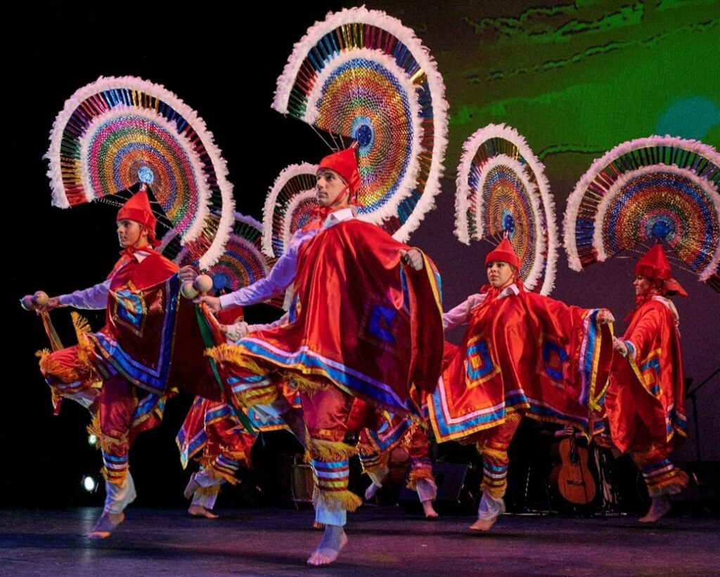 Color photo of dancers dressed in bright cultural costumes with large feathered head pieces dancing onstage.