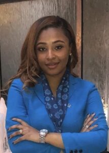 Mari Vieira-Gunn is a Black woman with long hair. She wears a bright blue jacket, printed blouse, watch and other jewelry. She is shot from the chest up with her arms crossed in front of her and a wooden wall behind.
