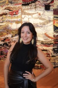 Fabiola R. Delgado is a Latino woman with long hair who wears gold feather shaped drop earrings, a blck leather skirt, and a black tank top. She is photographed from the waist up in front of a brightly colored weaving.