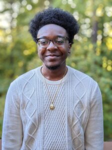 Jasir Qiydaar is a Black man with horn-rimmed glasses and a medium afro. He wears a cream cable knit sweater and a gold necklace. He is shot from the chest up outside in front of green trees.