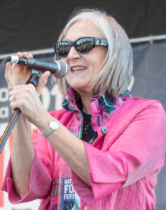 Color photo of Julia Olin standing behind a microphone on a stage. She is wearing a bright pink blouse and sunglasses.