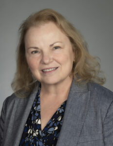 Headshot of Theresa Colvin. Colvin is a white woman with shoulder length auburn hair. She wears a black, white, and blue patterned blouse and grey blazer.