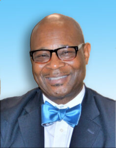 P. Muzi Branch is a Black man with a clean-shaven head and glasses. He wears a suit jacket, white collared shirt and bright blue bow-tie. He is photographed from the shoulders up in front of a blue background.