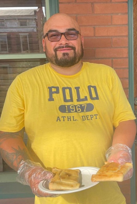 Sergio Herrera is shot from the waist up in front of a brick building with a glass door. He holds a white plate containing traditional Chilean food. He wears a bright yellow Tshirt has a dark short beard and wears glasses.