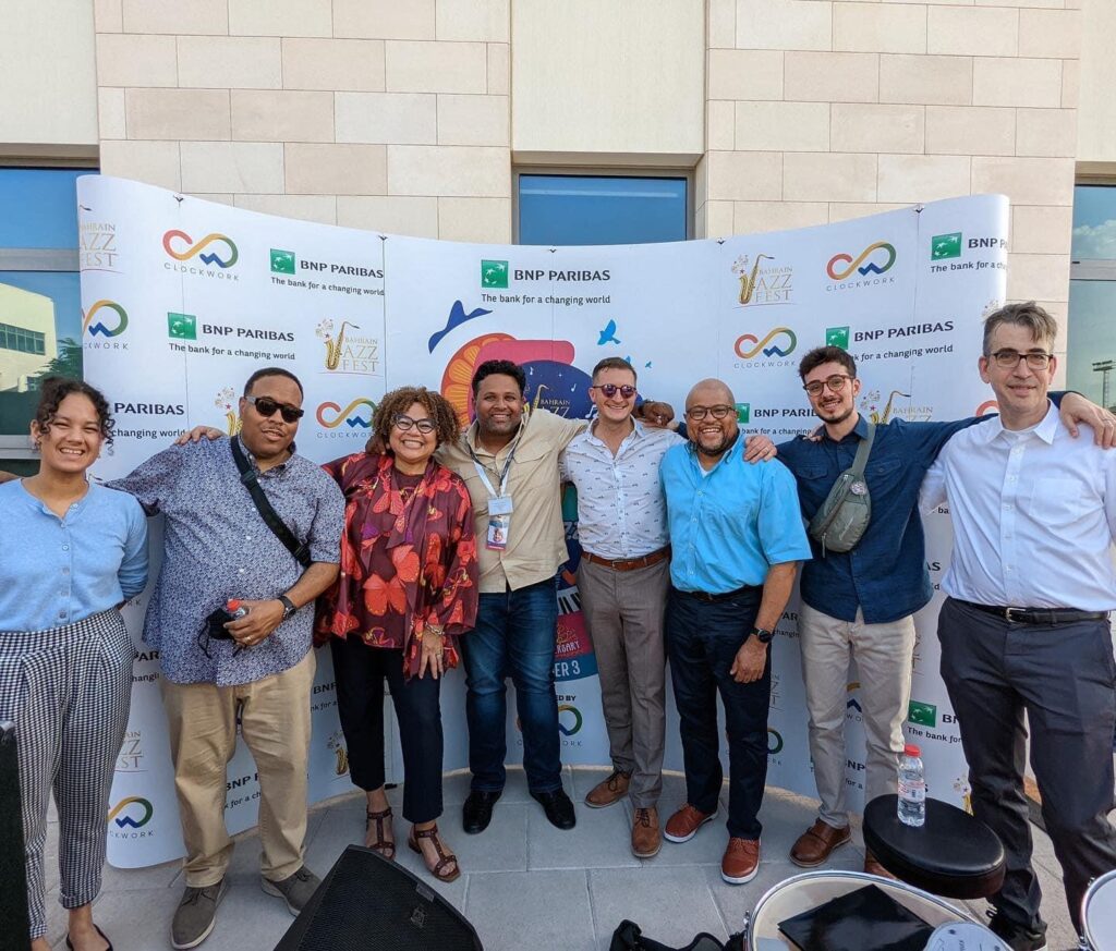 Eight members of the Jazz House Collective stand outside of a venue in front of a Bahrain Jazz Festival step and repeat backdrop. They are all dressed casually and have their arms around each others shoulders.
