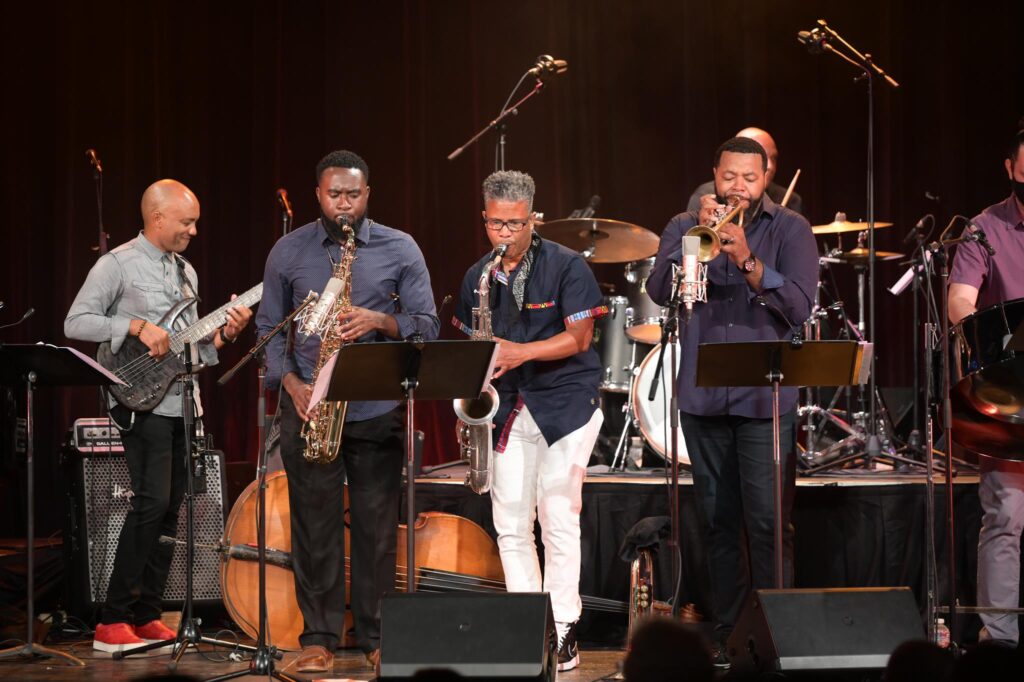 Four jazz musicians line the front of the stage. They are all playing their instruments - guitar, saxophone, trumpet. In the back, a drum set and percussionist can be seen.