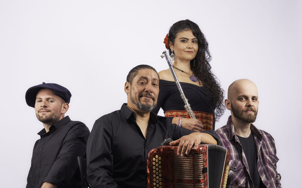 Three males musicians site on boxes in a semicircle. They all ear shades of black with accents. To the left a bass lays on the floor. The center musician holds an accordion. The man to the right has an acoustic guitar. Behind them standing center is a woman with a flute.