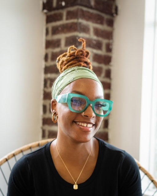 Femeika Elliott is shot from the chest up. She sits in a chair in front of white walls and a brick corner piece. She wears a black shirt and a gold necklace and earrings. She wears dreads in a bun and turquoise framed glasses.