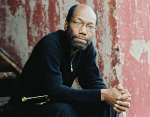 Charles Tolliver sits on concrete steps with his trumpet across his lap/ Behind him is a concrete wall with brick red paint peeling off. Mr. Tolliver is an African American man with a beard and glasses.