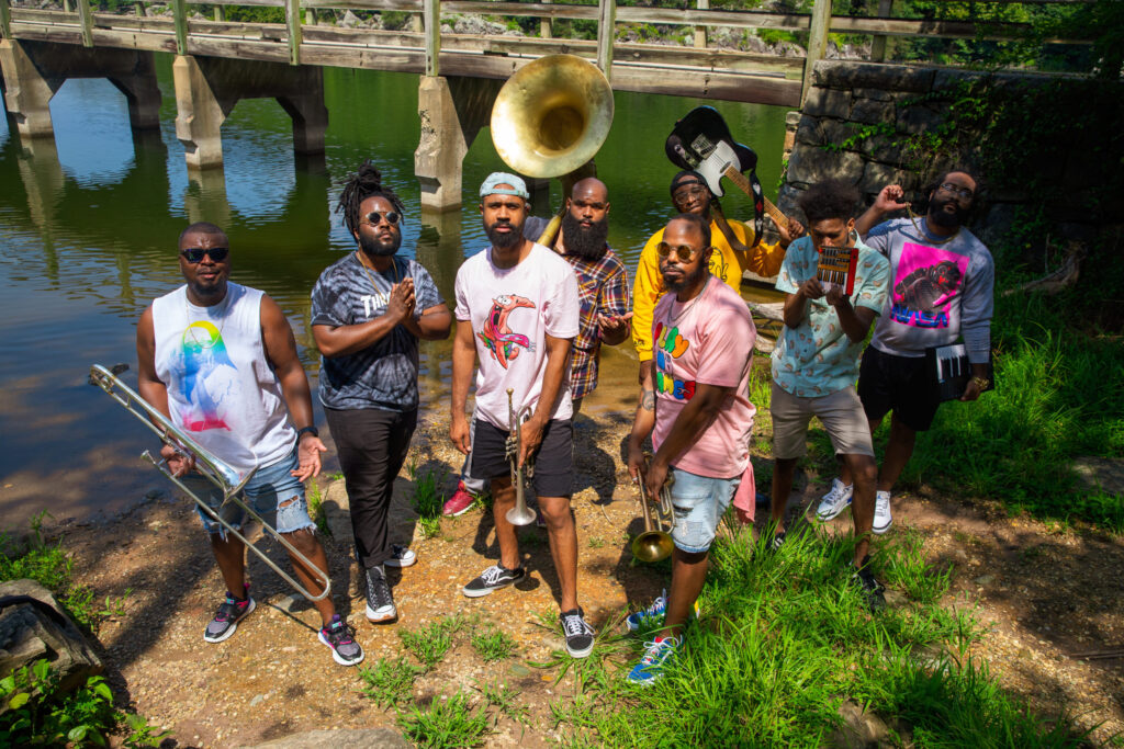 A group of eight musicians in casual summer clothing stand on the banks of a river with their instruments in their hands. Behind them, a wooden bridge can be seen.