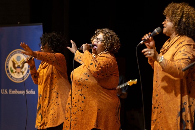 Color photo of three women in matching long orange tops singing onstage with handheld mikes. A guitarist is visible in the background.