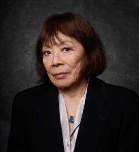 Head shot of Toshiko Akiyoshi. Toshiko is an Asian woman with brown hair cut into a chin length bob with bangs. Her head is turned slightly to the right. She is wearing a black blazer with a soft white v-neck blouse and necklace.