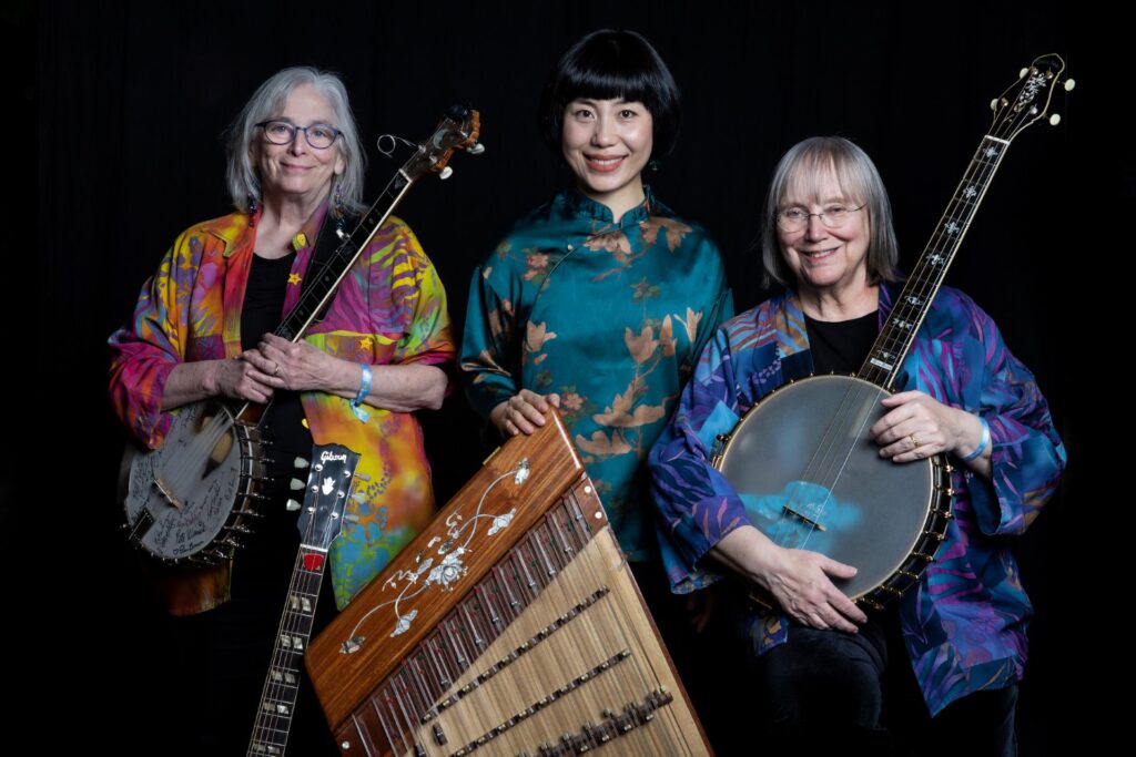Three women stand against a dark background. They are dressed in vibrant clothing. They all hold stringed instruments and a traditional Chinese wooden instrument sits across the front of the group.