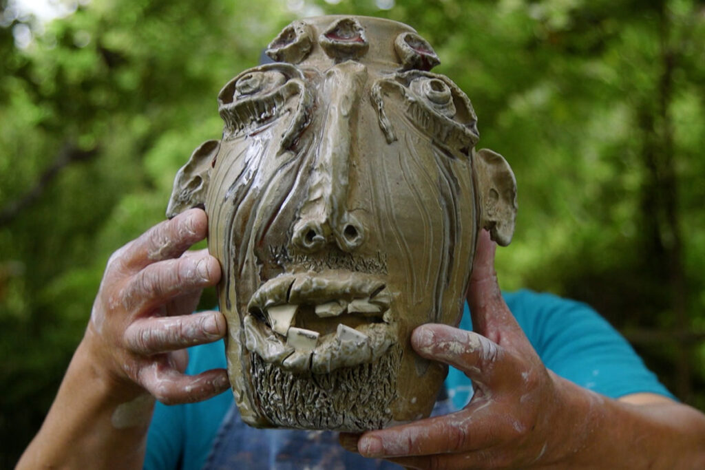 Color photo of a man holding a clay jug head sculpture in front of him.
