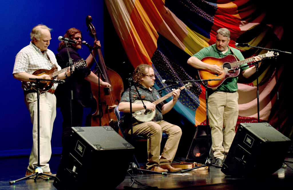 Four men perform on mandolin, banjo, upright bass, and guitar in front of a quilted backdrop