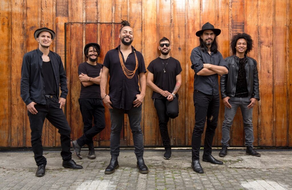 Color photo of six musicians dressed in black posed in front of a wide board wood wall.