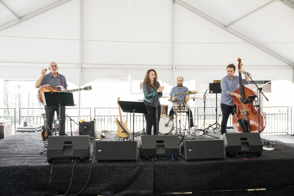 A four piece band performs on a riser stage under a white pavilion.