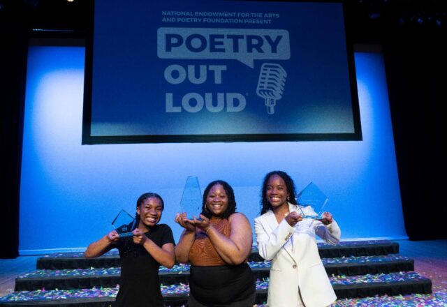 Three people stand center stage holding glass trophies. Behind them, a screen with the words Poetry Out Loud can be seen.