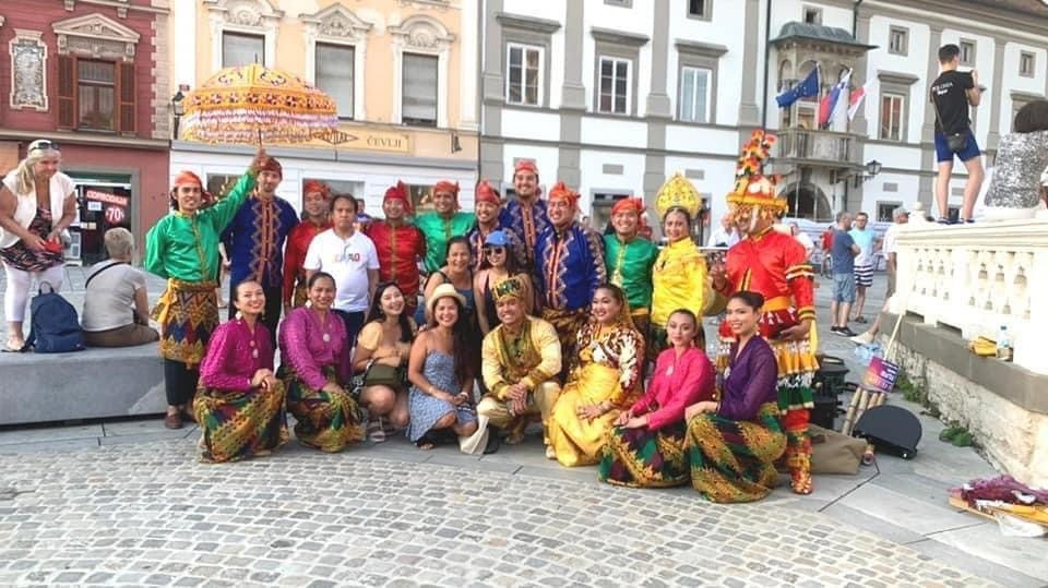 A group of brightly dressed people pose on a town square for a photo.
