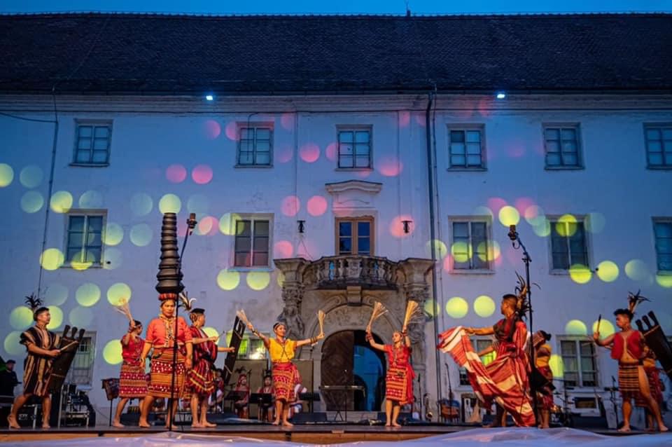 A group of dancers in traditional costumes perform on a stage in front of a brightly lit building.