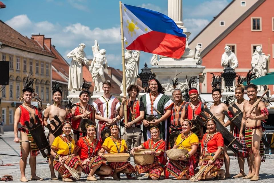 A group of people in traditional dress pose for a group photos outside in a city street. A flag flies above them.