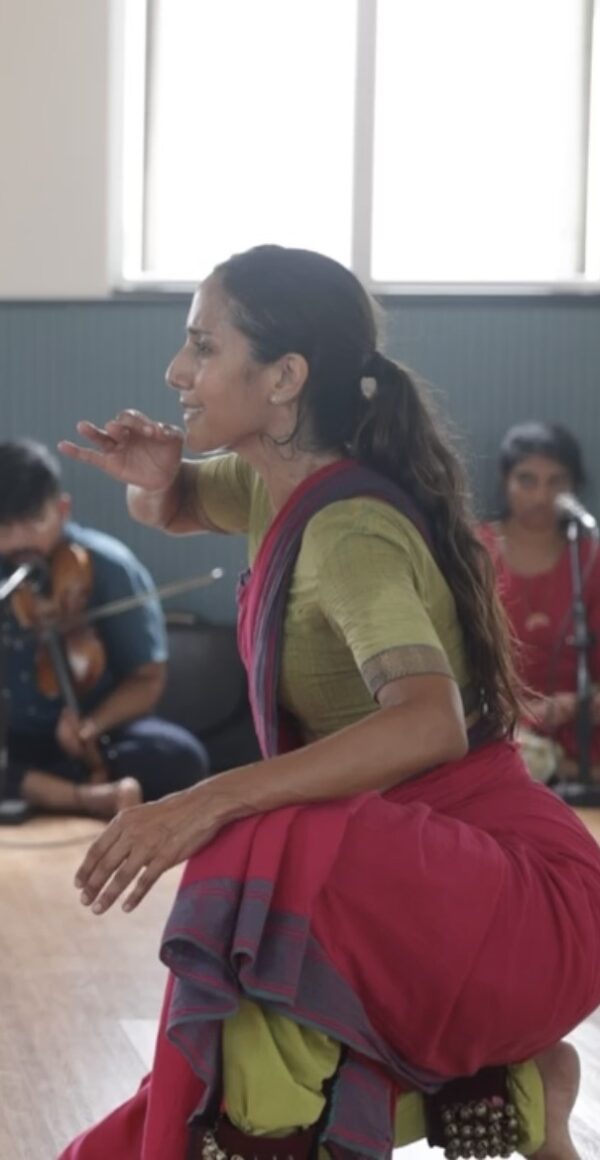 A dancer in a brightly colored sari crouches in profile to the right.
