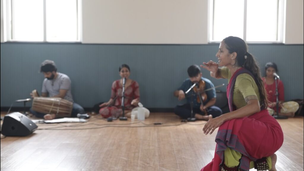 A dancer in a colorful sari kneels in profile on the right. Behind her musicians can be seen against a studio wall.