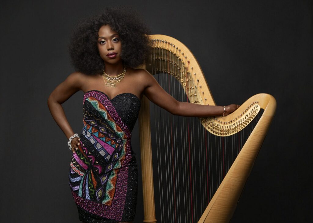 Brandee Younger wears a bright dress and green shoes while standing beside a tall gold harp one hand draped over the top of the instrument.