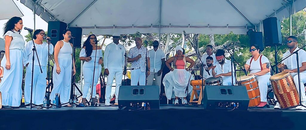 A large group of performers dressed in white line an outdoor stage. Some sing into microphones while others play traditional instruments.