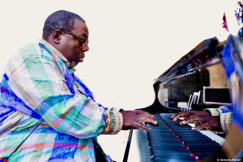 Cyrus Chestnut is seated and playing piano, wearing a brightly colored pastel plaid shirt. His hands are reflected in the shining piano fallboard.