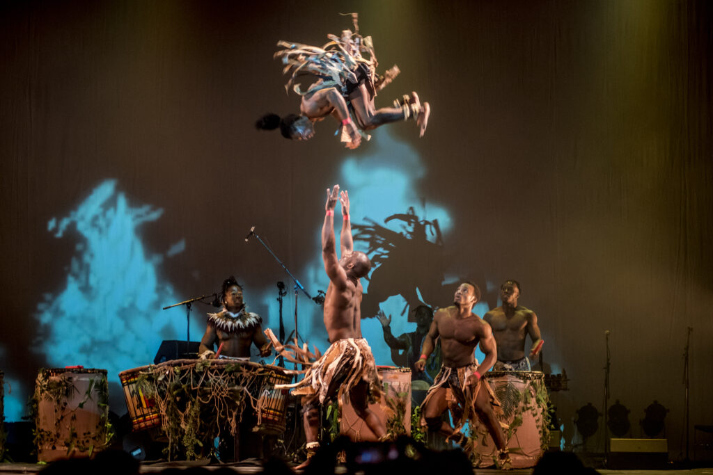 A man throws a fellow acrobat in a flip in the air while drummers in the background look on. The performers are all in authentic African costumes.