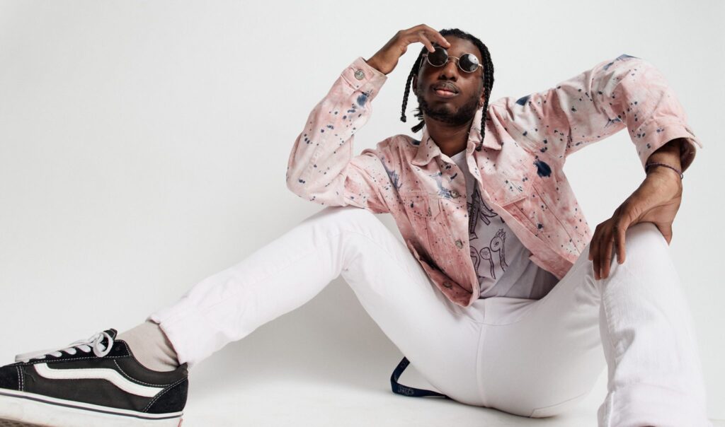 Julius Rodriguez is shot in front of a white background. He wears white pants, black sneakers and a pink patterned jacket with sunglasses.