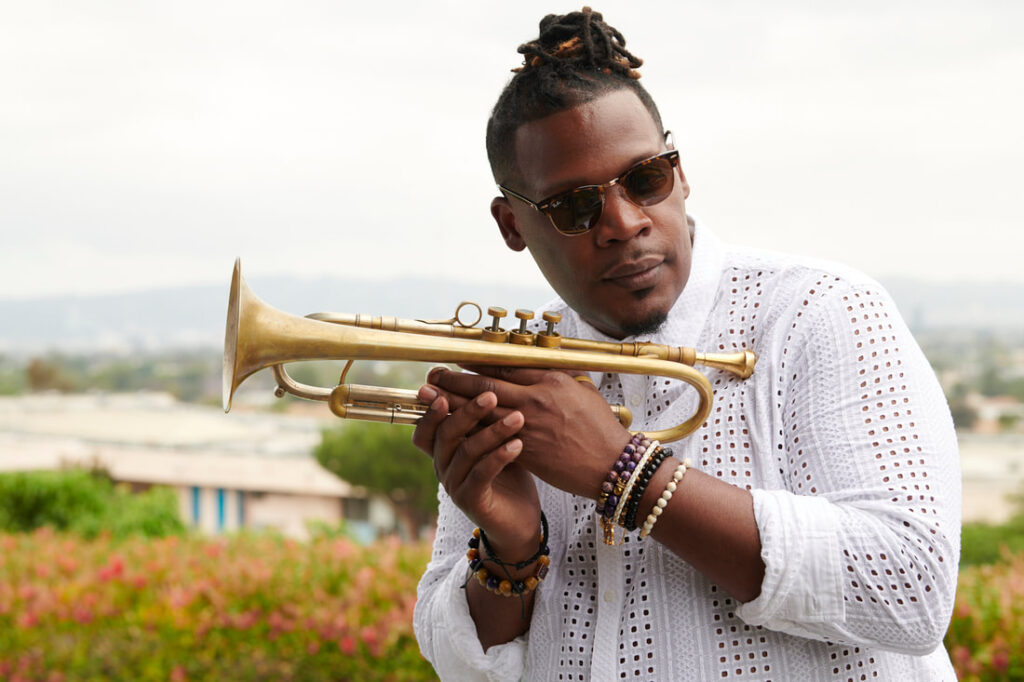 Keyon Harrold stands in a field holding a trumpet. He wears a white shirt and sunglasses.