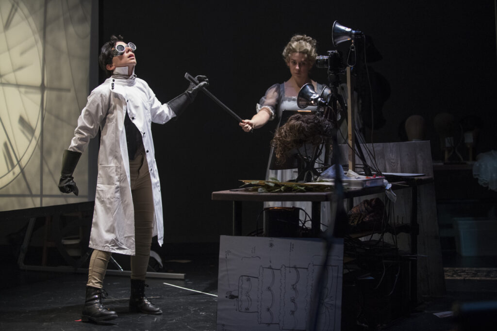 Two actors appear on a stage. One wears a lab coat and goggles while the other hands them a tool. A projection is visible behind them.