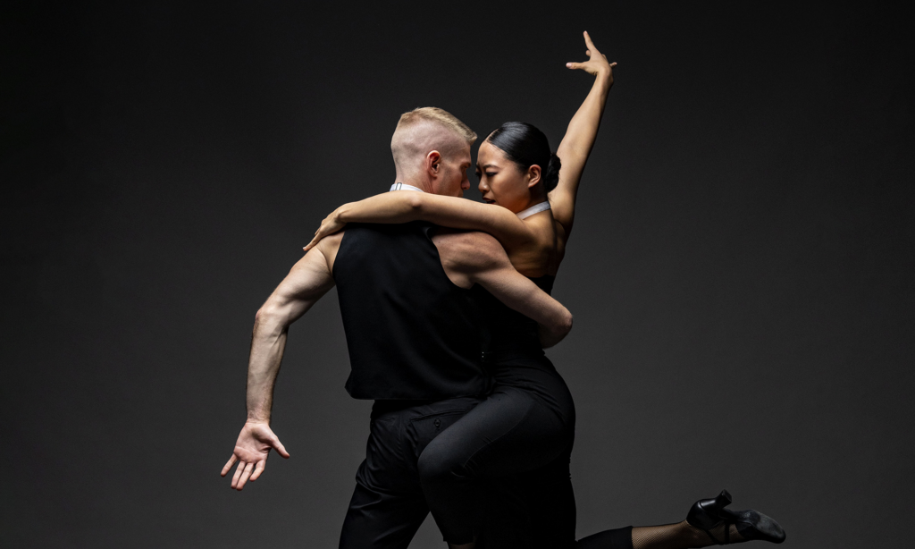 Two dancers in black outfits perform a dramatic dance pose, with one dancer lifting the other.