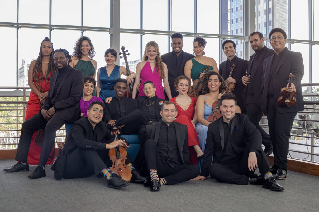 A large group of people dressed in formal wear pose inside of a modern building with floor to ceiling windows behind them. Some of the people hold stringed instruments.