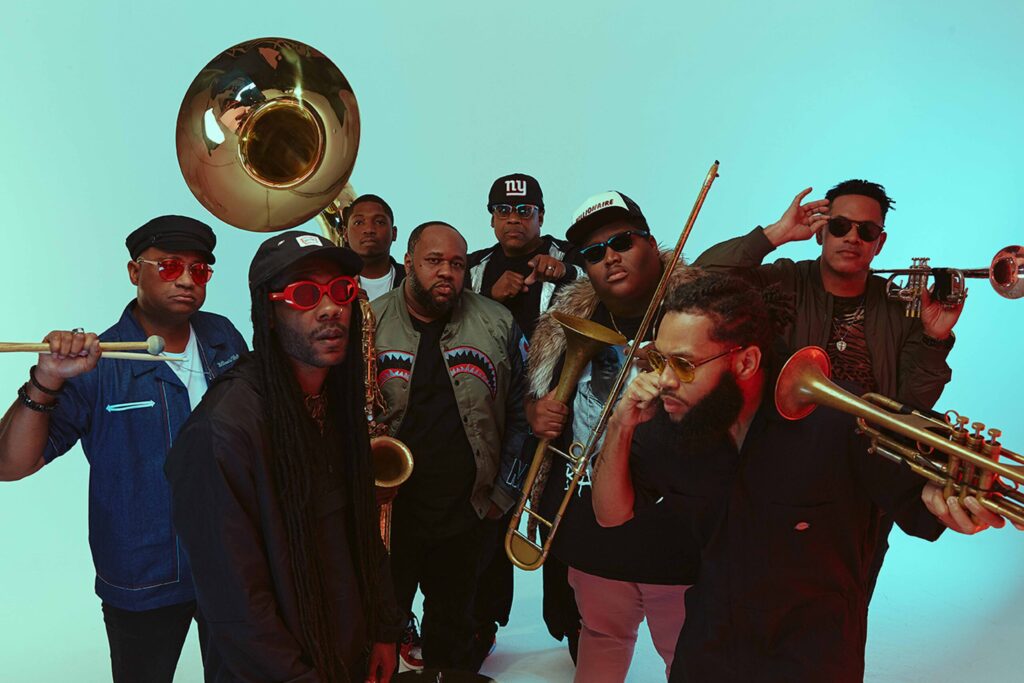Studio shot against a blue background of eight musicians with their instruments in hand.