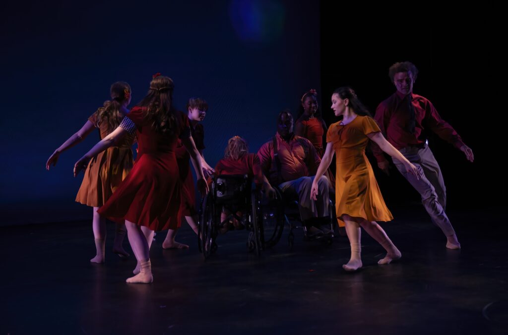 Six dancers gracefully encircle two performers in wheelchairs on a dimly lit stage. Dressed in warm hues red and orange, Dancing Wheels formed a mesmerizing dance, blending inclusivity and artistry into a poignant spectacle of unity and diversity.