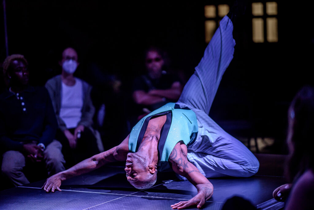 A dancer arches back on the catwalk one knee on the floor and both hands over their head behind them. One leg point up to the ceiling.