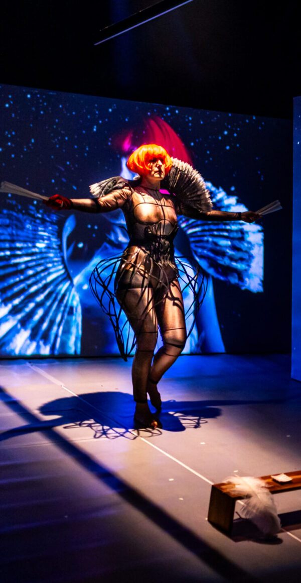 A performer walks down a dark runway on stage. They wear a dark futuristic jumpsuit. A projection of them is behind and the stage is bather in blue and white.