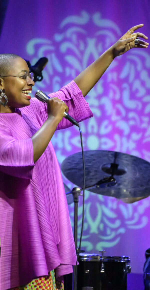 A vocalist raises their left hand in the air while singing into a mic in their tight hand. They wear a bright pink top and the stage is bathed in purple and blue light.