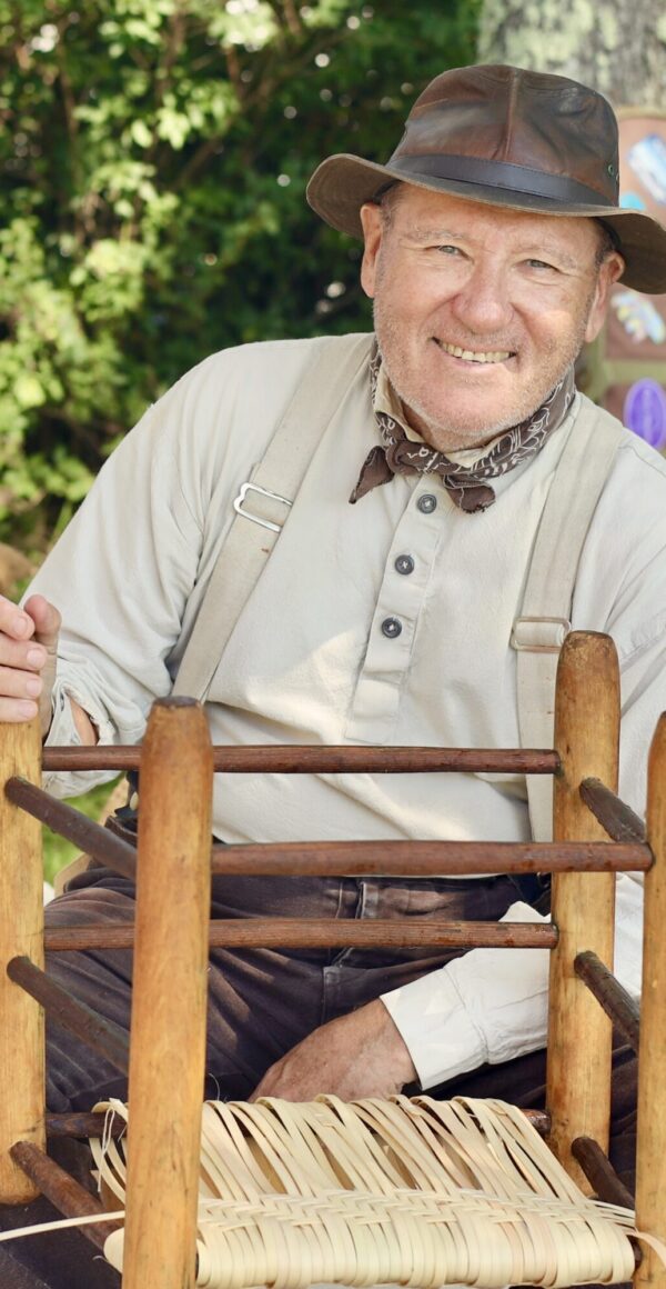 An artist works on weaving a chair seat while at an outside demonstration.